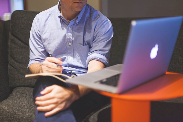 Muž v modrej košeli sedí na pohovke s zošitom na kolenách a pozerá o počítača.jpg